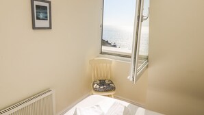 Ein Schlafzimmer mit Bett, Sessel, Heizkörper, Fenster mit Meerblick und einem gerahmten Bild an der Wand.