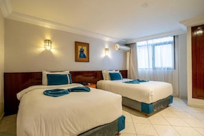 Une chambre d’hôtel avec deux lits, une tête de lit en bois, un climatiseur fixé au mur et une fenêtre avec des rideaux.