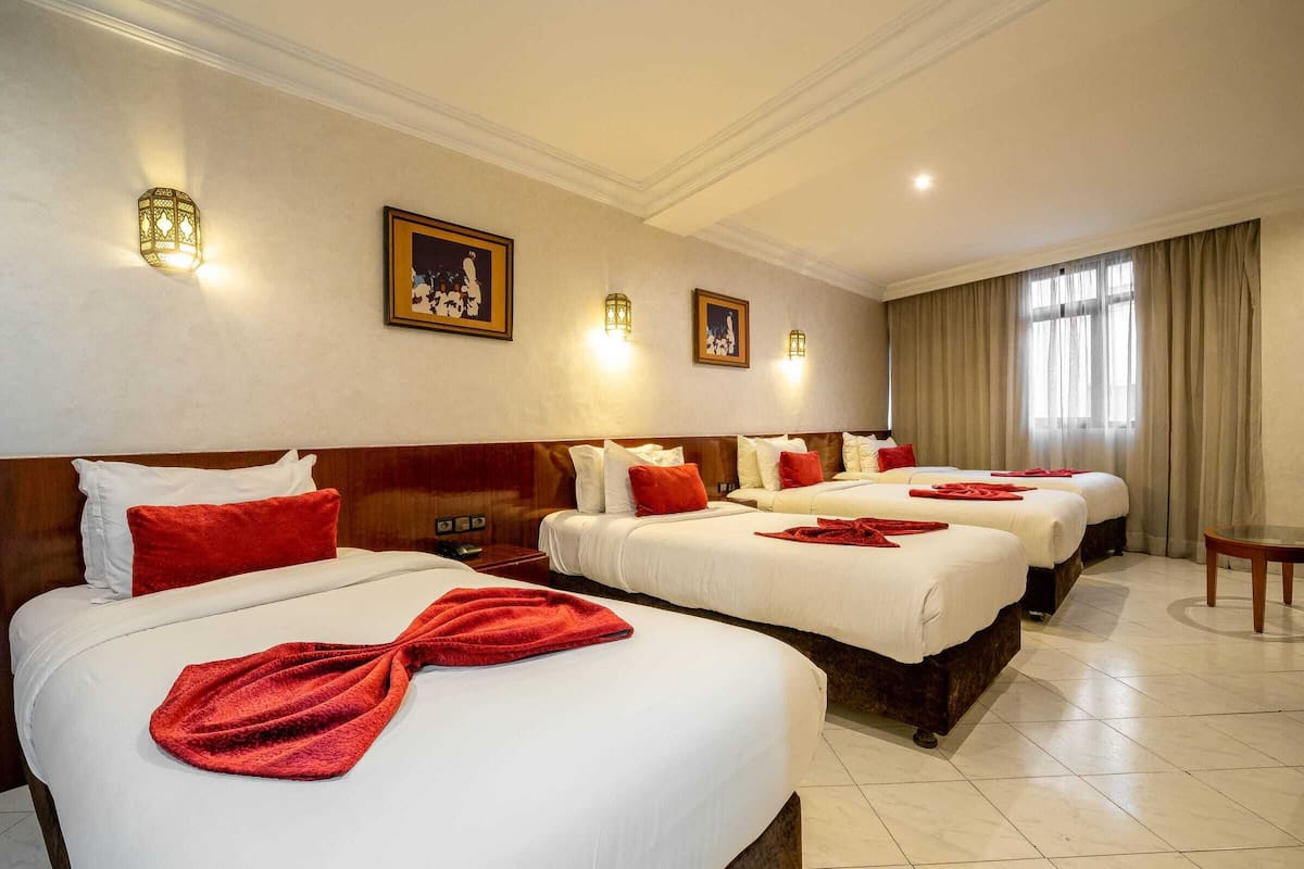 Habitación de hotel con tres camas, cada una con ropa blanca y almohadas rojas. Hay luces fijadas a la pared y cuadros enmarcados.