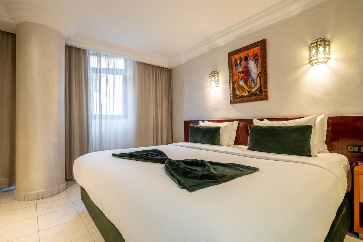 Habitación de hotel con una cama grande, cabecero de madera, dos lámparas de pared, un cuadro y una ventana con cortinas.