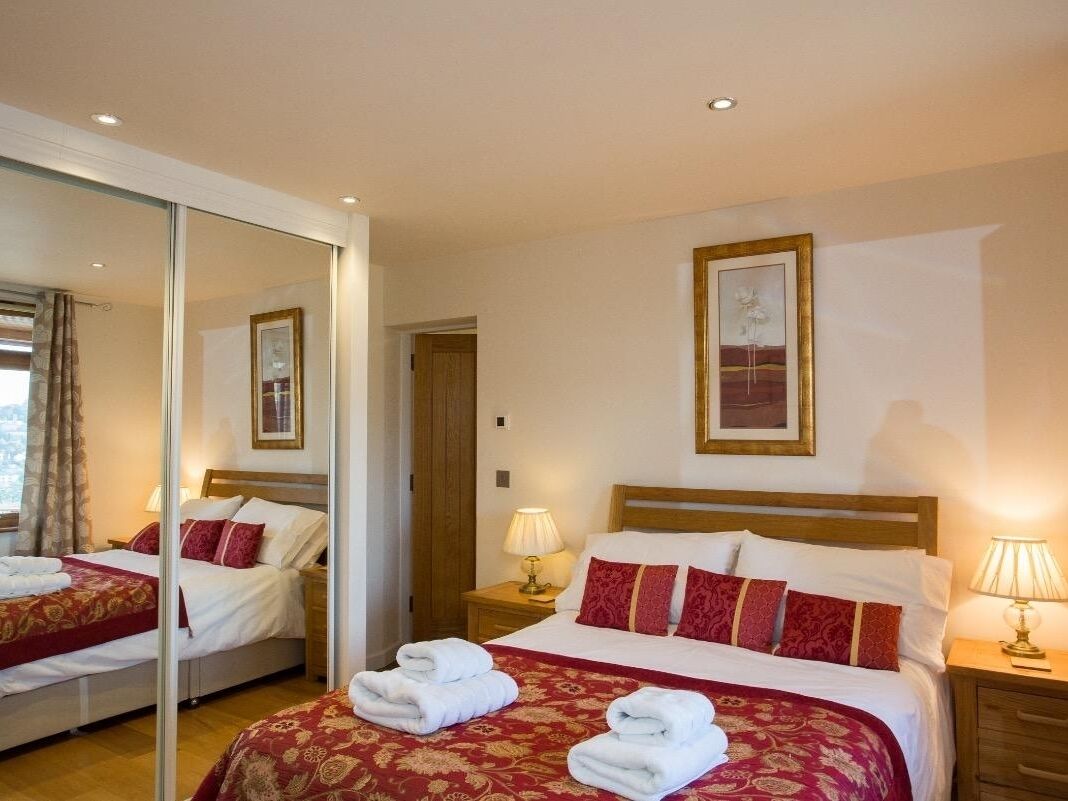 Habitación de hotel con un espejo grande, dos camas con ropa de cama roja y blanca, un cabecero de madera y lámparas de noche.