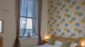 Un dormitorio con cama, ventana con cortinas, mesita de noche y papel tapiz floral en la pared.