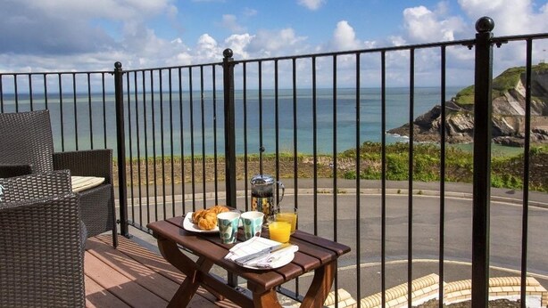 Un balcón con mobiliario de mimbre, una mesa de madera con provisiones para el desayuno, y vistas al mar y a los acantilados.