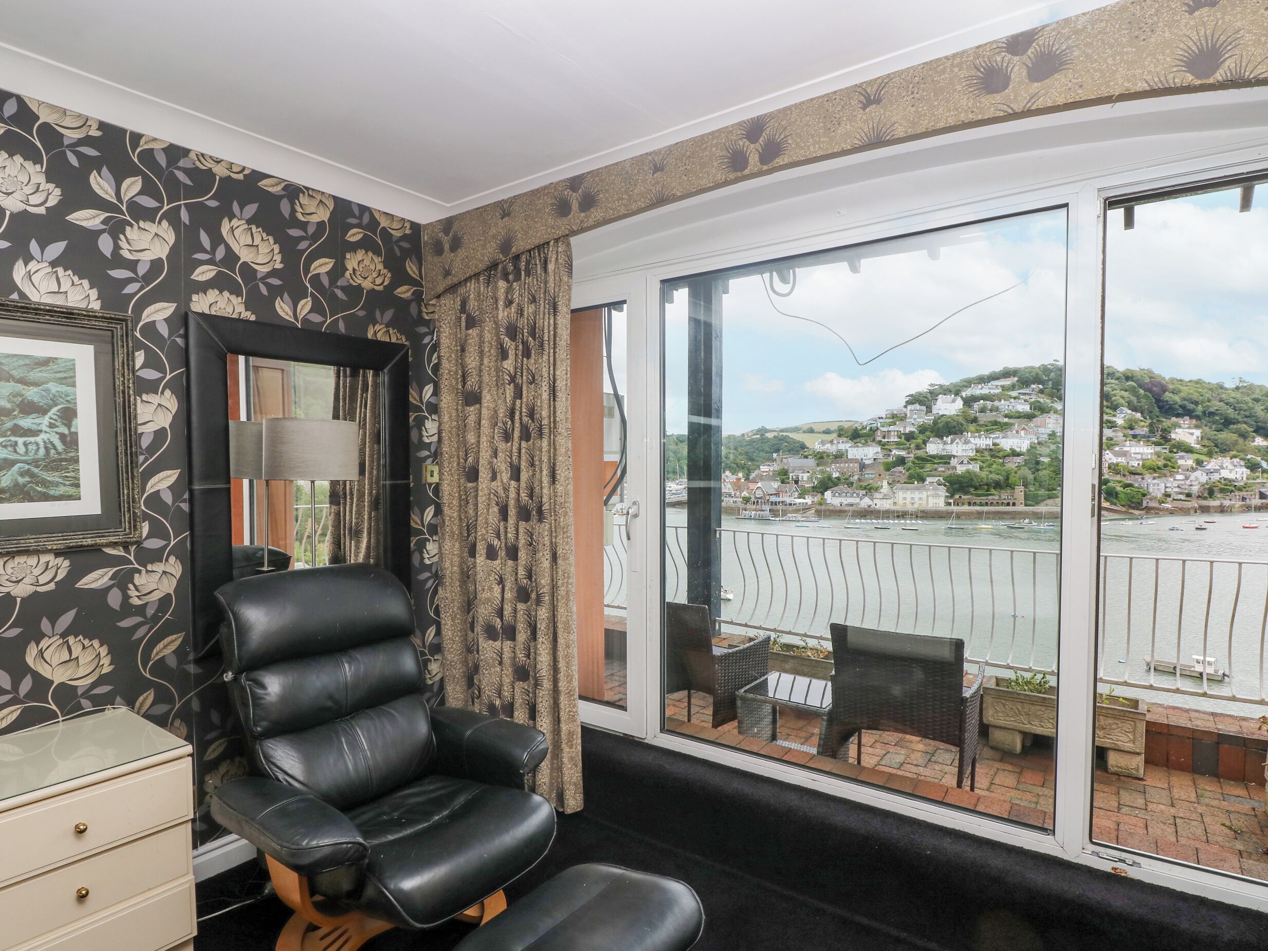 A room with a black leather chair, a framed picture on the wall, and a large window with a view of a harbor and buildings.
