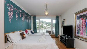 A hotel room with two beds, a TV, a painting, and a view of a city through the window. - Bight Boathouse (Dartmouth)