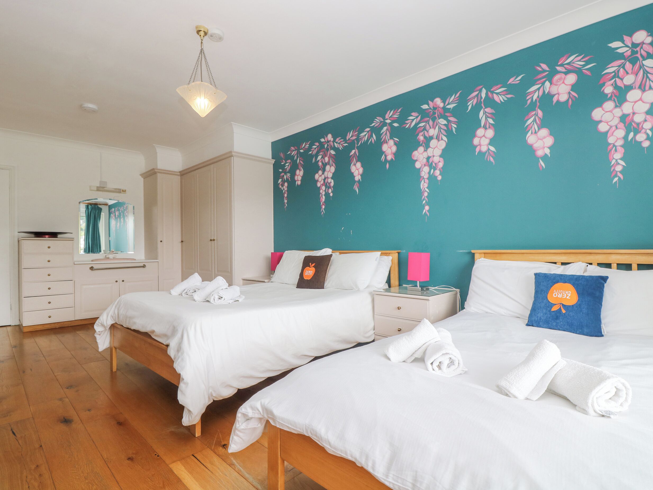 Una habitación de hotel con dos camas, un mural de pared color turquesa, suelo de madera, un aparador blanco y un espejo.