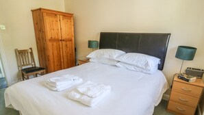 Un lit bien fait, avec du linge de lit blanc et des serviettes pliées, une armoire en bois, une chaise et une table de chevet avec une lampe et des livres.