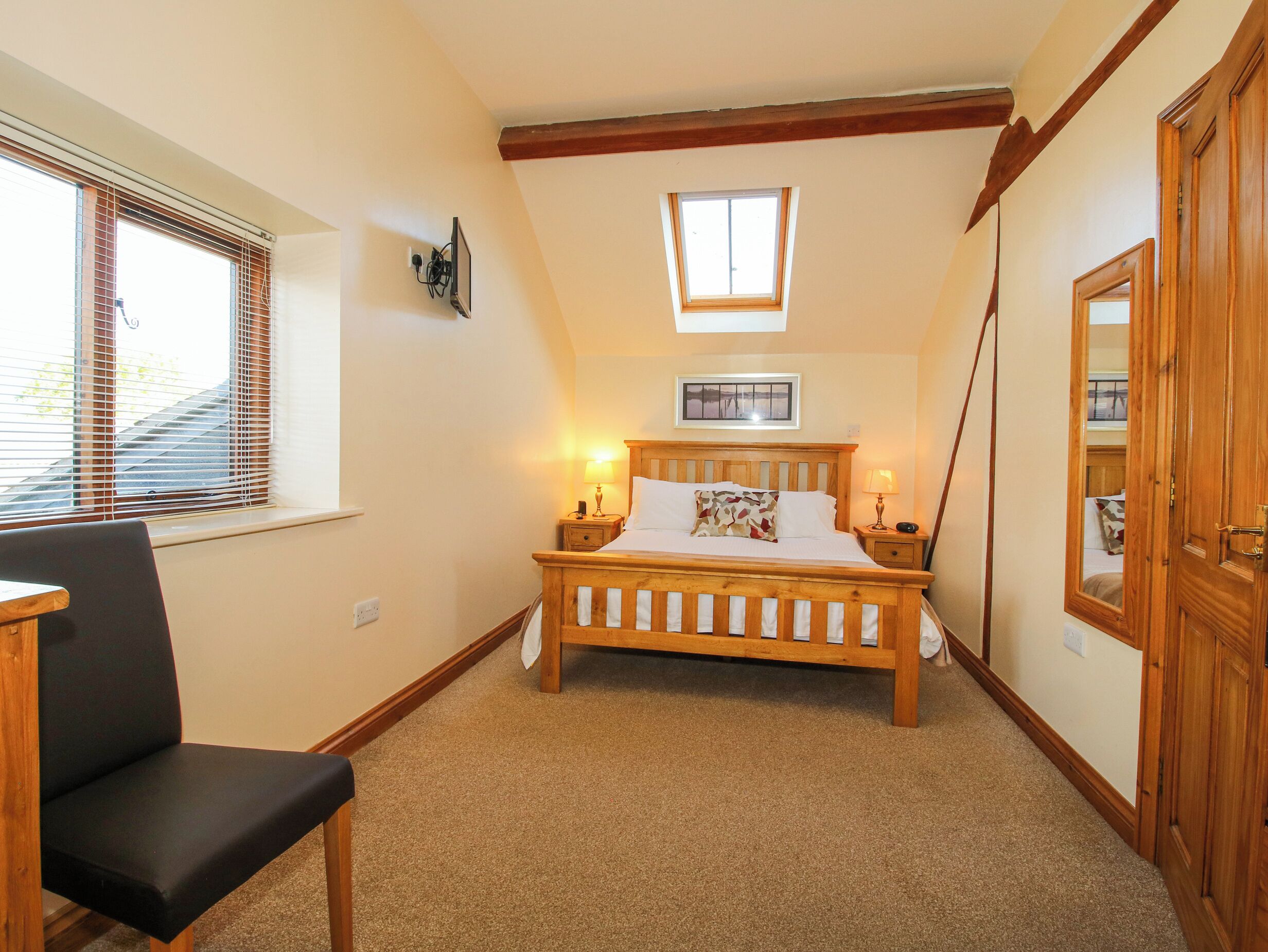 A bedroom with a wooden bed, a chair, a window with blinds, a small skylight, and a door.
