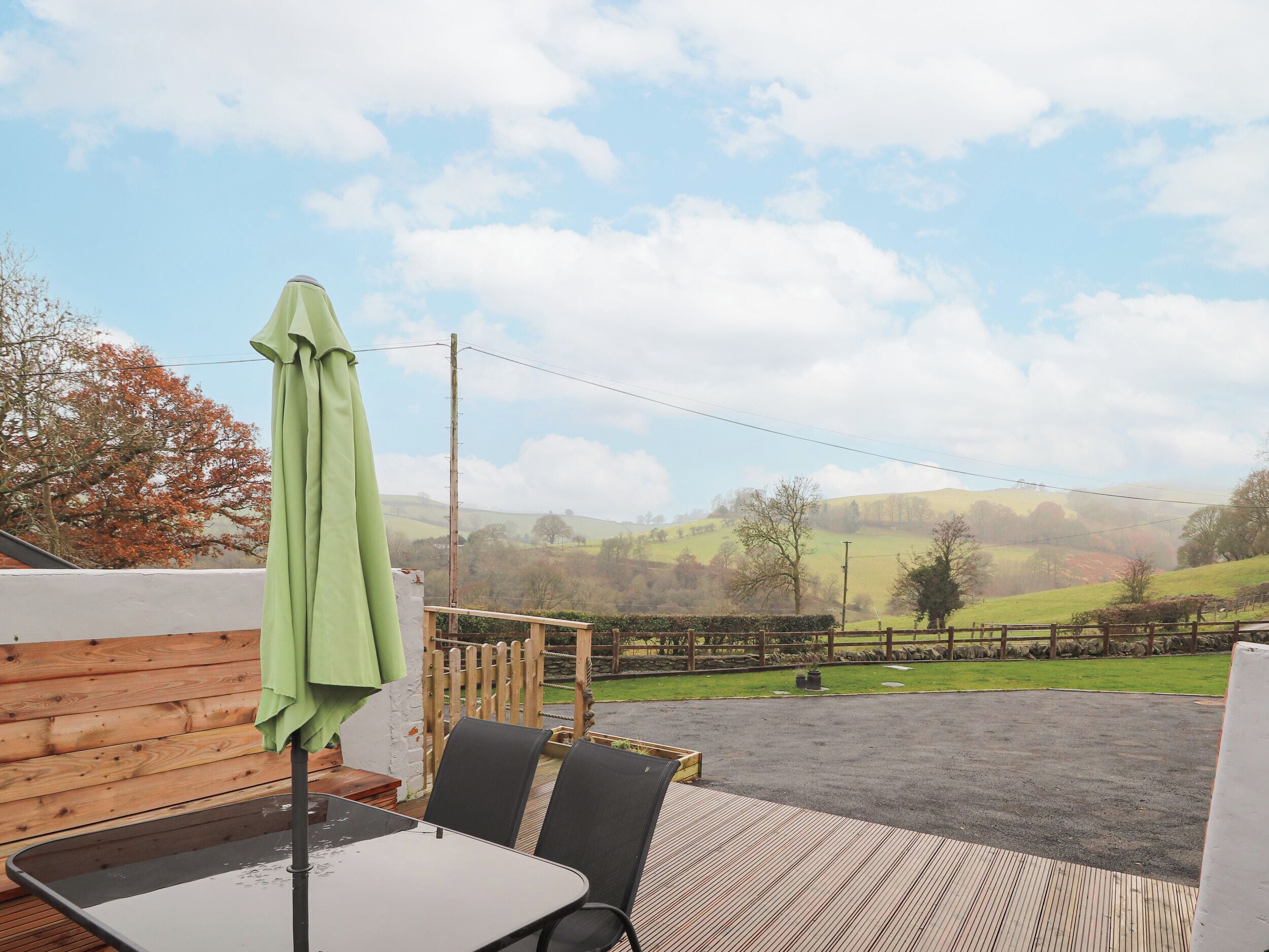 A patio with a table and chairs, a green umbrella, and a view of a rural landscape with hills and trees.