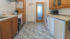 A compact kitchen with wooden cabinets, a built-in oven, a washing machine, and a microwave. - Salty Puffin (Tyn-y-Gongl)