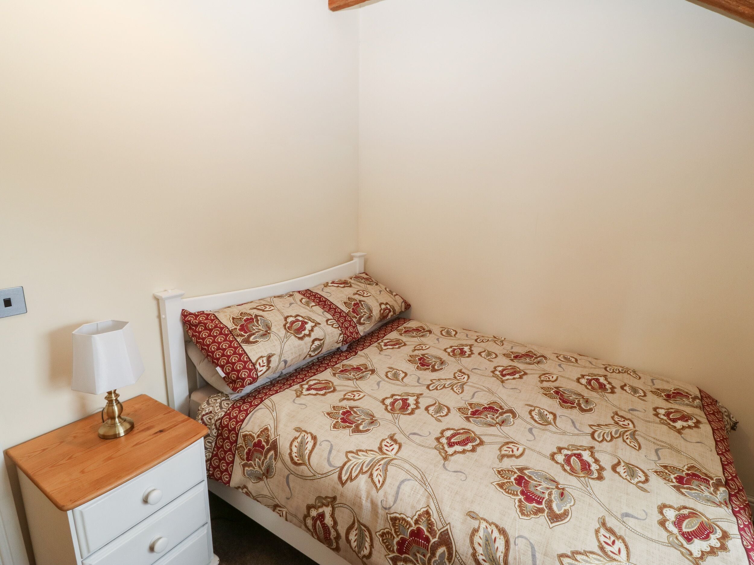 Une petite chambre avec un lit simple, une table de chevet en bois, une lampe blanche et une couvre-lit à motifs.
