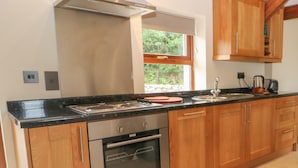 A modern kitchen with wooden cabinets, stainless steel appliances, and a large window providing natural light. - Y Beudy Cottage (Caernarfon)