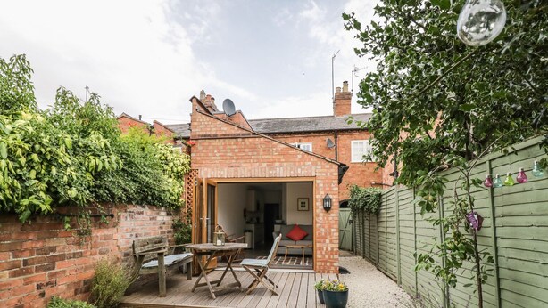 A brick patio with a wooden table and chairs, a small garden area with gravel paths, and a green fence. - 24 College Lane (Stratford-upon-Avon)
