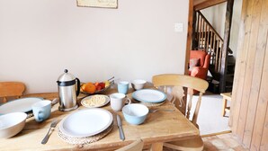 Un comedor con una mesa de madera dispuesta para cuatro personas, con platos, tazones y cubiertos. Se ve también una cafetera y un frutero.