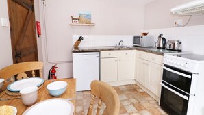 Cocina compacta con alacenas blancas, un microondas, un tostador y un lavavajillas. Hay una mesa de comedor de madera con platos y tazones, y una silla. Un extintor está montado en la pared.