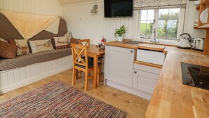 A cozy cabin interior with a wooden dining table, a sofa with patterned cushions, a television mounted above the table, and a window with a view of greenery. - Hillside View (Rhayader)