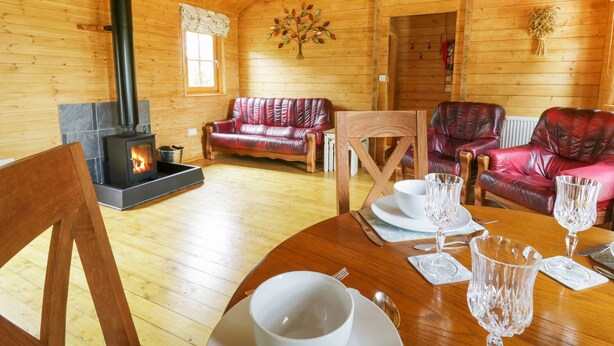 A cozy cabin interior with a fireplace, leather sofas, a dining table set for two, and wooden walls. - Acorn Lodge (Saxmundham)