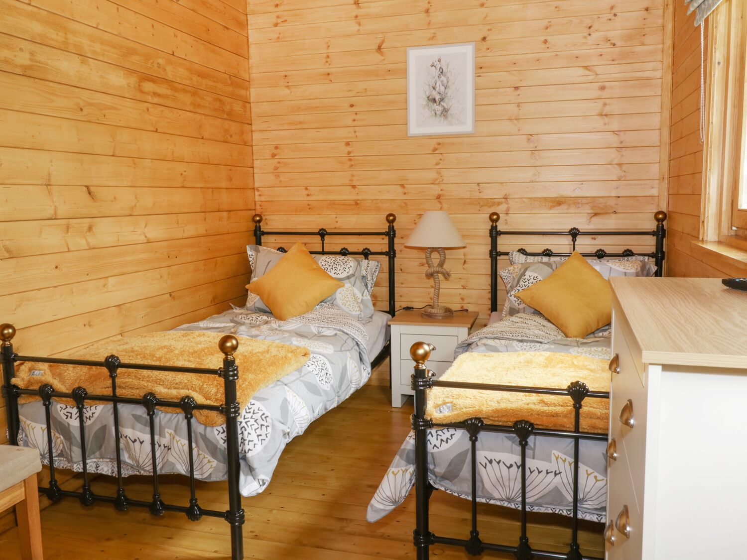 Quarto de cabana de madeira com duas camas, uma mesa de cabeceira com abajur e um quadro na parede.