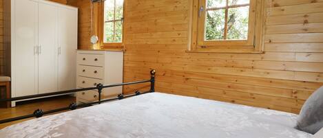 A wooden cabin room with a bed, a wardrobe, a dresser, and two windows with curtains.