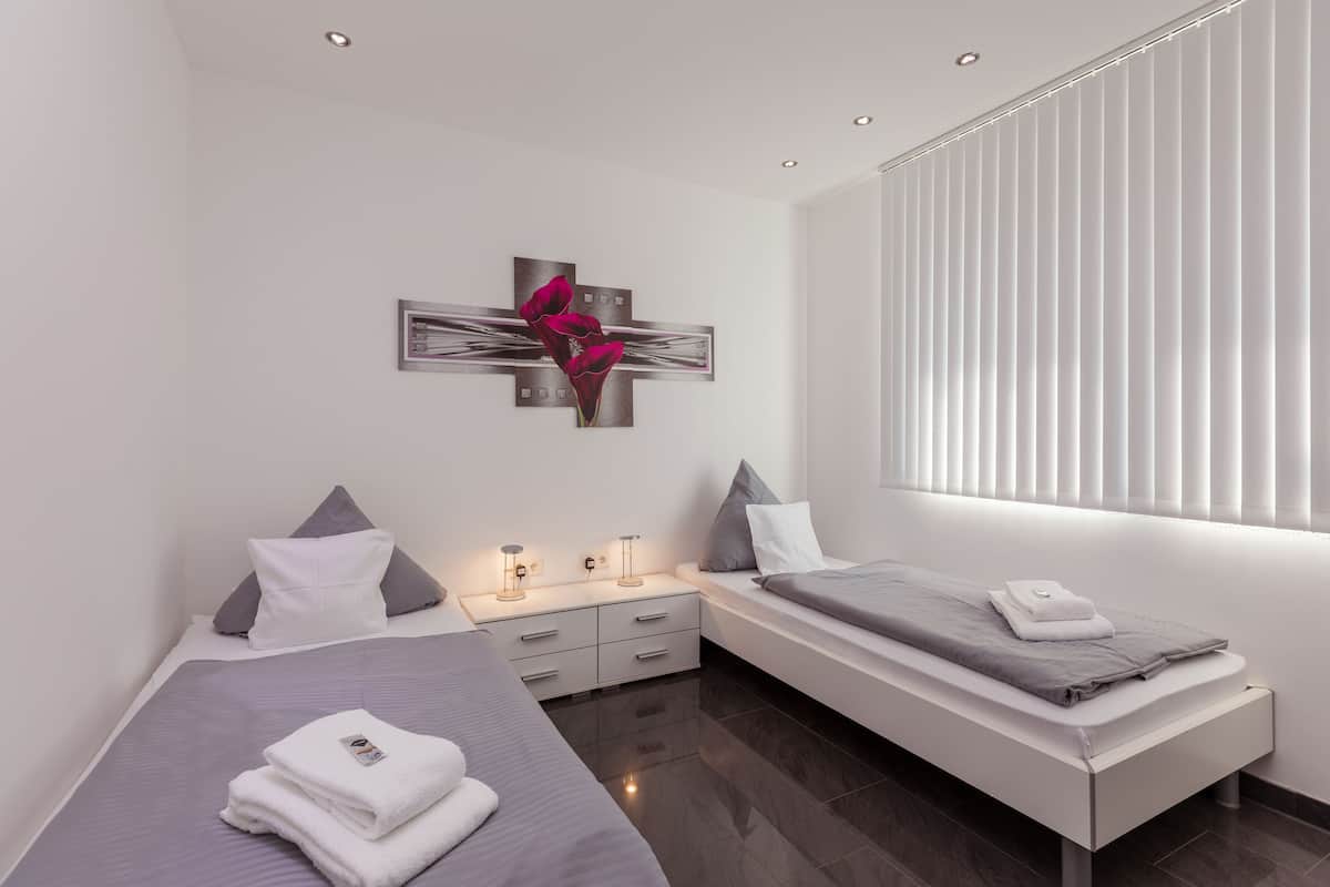 Une chambre moderne avec deux lits, une table de chevet, une œuvre d’art accrochée au mur et une fenêtre avec des stores.