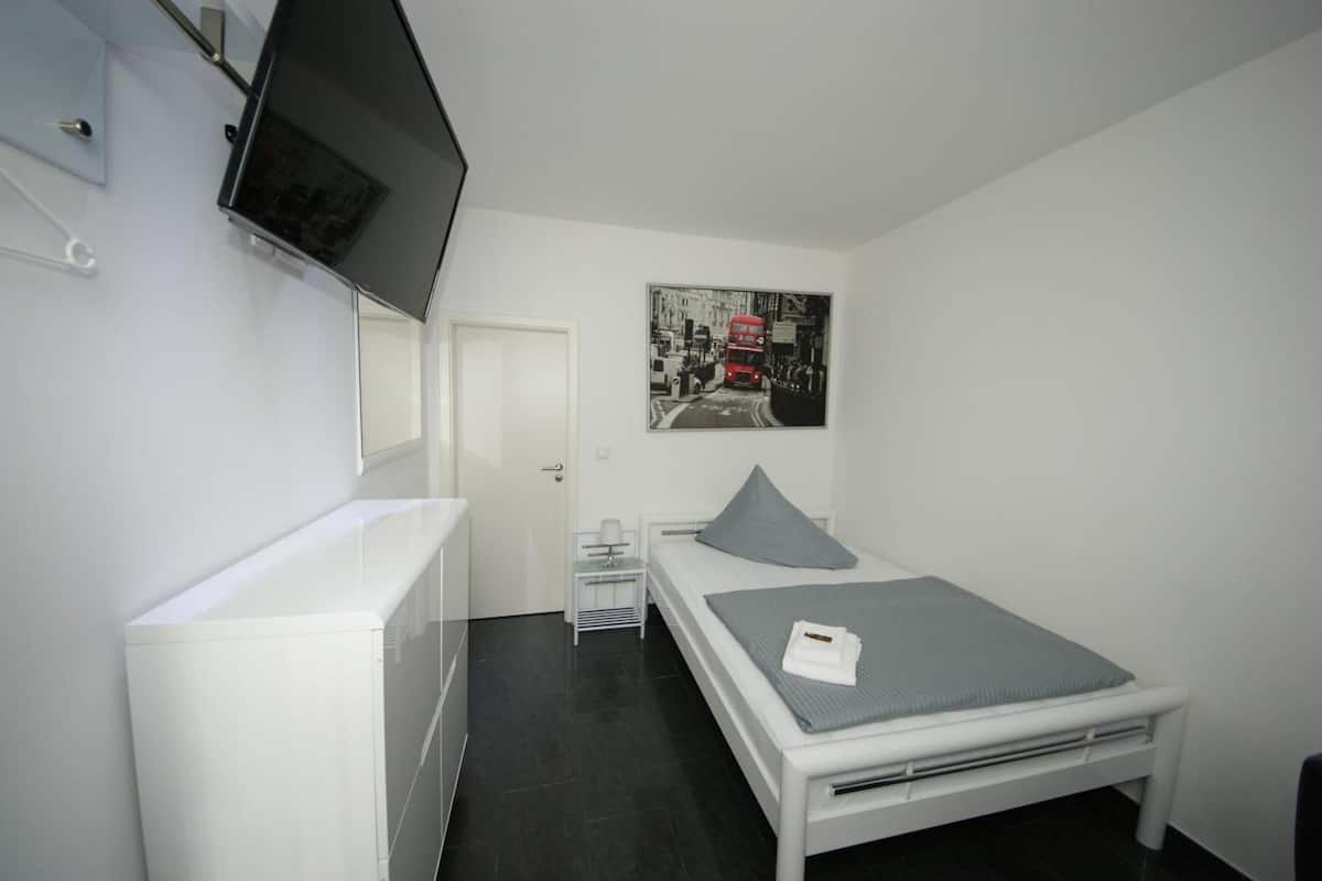 Une petite chambre moderne avec un lit simple, un téléviseur à écran plat fixé au mur, une petite table de chevet et une porte donnant sur une autre pièce.