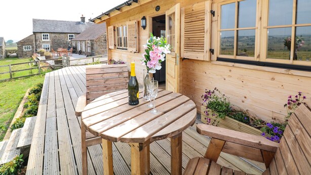 A wooden deck with a round table and benches, a bottle of champagne, and a vase of flowers. - The Shooting Lodge (Buxton)