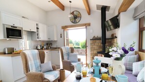 A cozy kitchen with white cabinets, a built-in microwave, and a stone fireplace. There are two upholstered chairs and a dining table set with breakfast items. - Sunnybrook Cottage (Clitheroe)