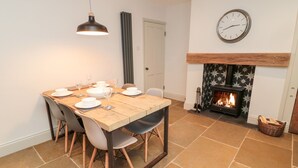 Un coin repas avec une table en bois dressée pour quatre personnes, un luminaire suspendu moderne, une cheminée ornée d’un motif en étoile et une horloge murale.