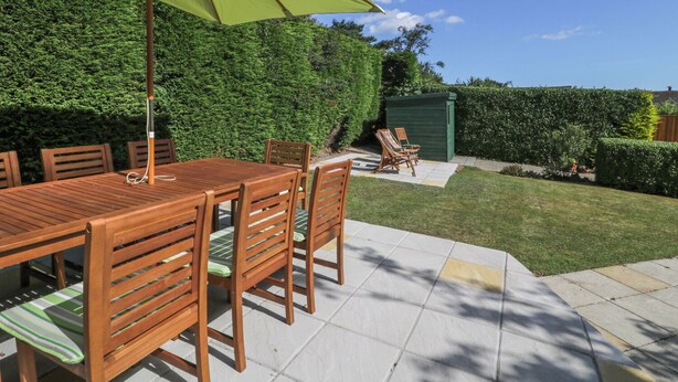 Une terrasse avec une table et des chaises en bois, un parasol vert et une haie en arrière-plan.