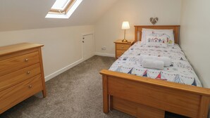 A bedroom with a sloped ceiling, a skylight, a bed with a nautical-themed duvet, a bedside table with a lamp, and a wooden dresser. - Bay View (Tyn-y-Gongl)