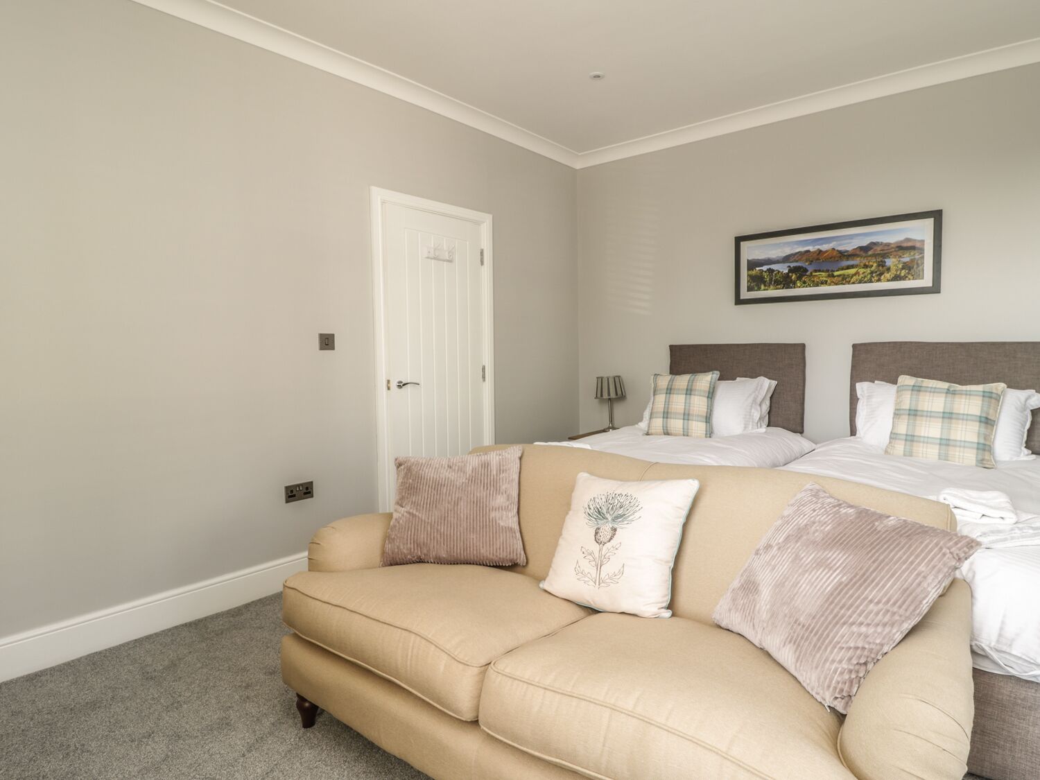 A bedroom with a beige sofa, two beds with matching headboards, a door, and a framed landscape picture on the wall.