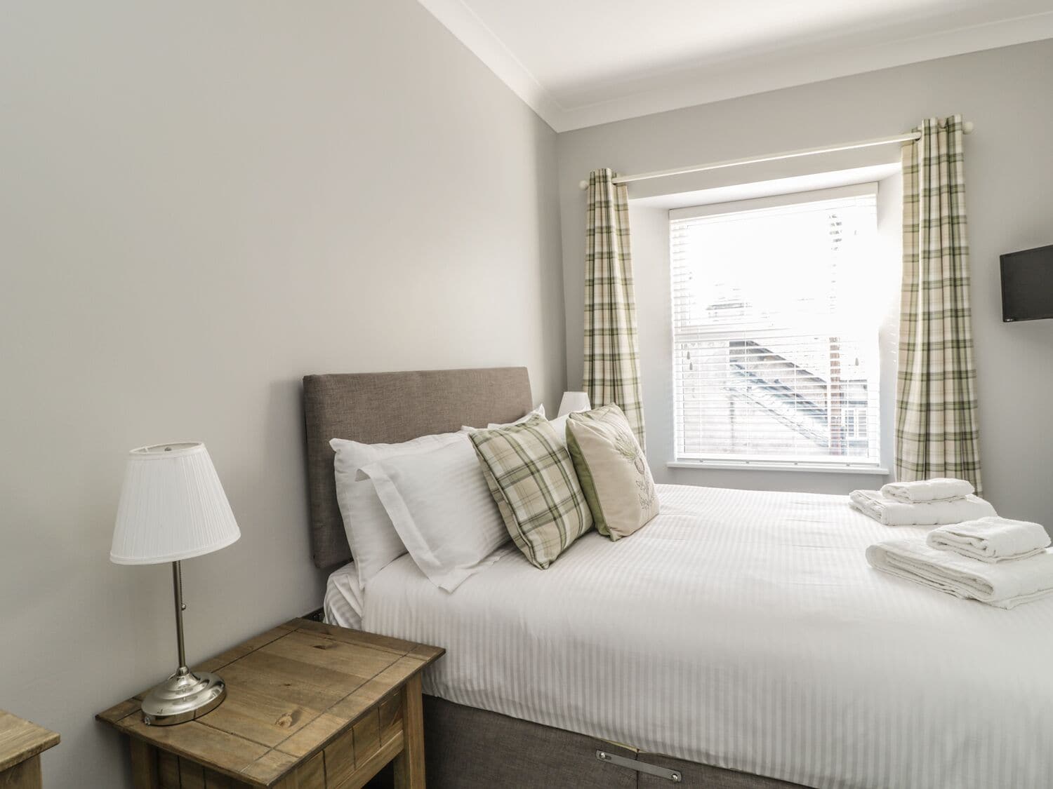 A neatly arranged hotel room with a bed, a bedside table with a lamp, a window with curtains, and a view of a building outside.