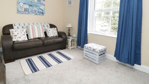 Un salón acogedor con un sofá de cuero marrón, alfombra a rayas y cojines con motivos náuticos. Una pequeña mesa con lámpara y ventana con cortina azul.