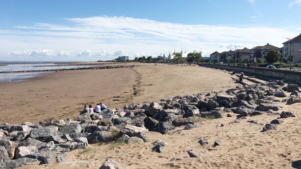 Una playa arenosa con rocas grandes al frente, cielo despejado y edificios en la distancia.