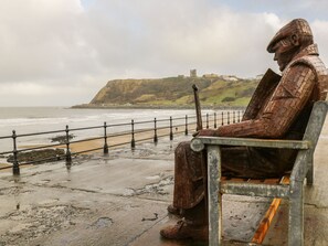 Une statue représentant une figure assise sur un banc, donnant sur une zone côtière, avec un bâtiment historique en arrière-plan.