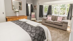 Una camera da letto con un letto grande, una cassettiera in legno, un divano grigio con cuscini rosa, una poltrona e una finestra con tende.