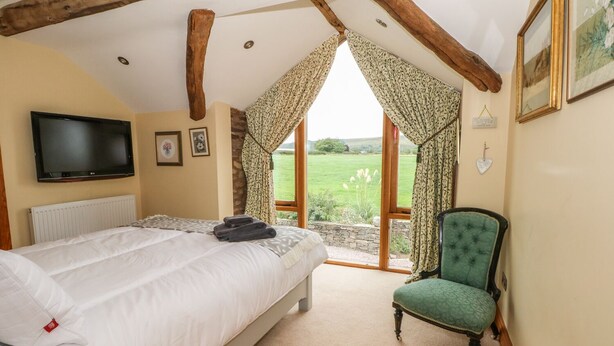 A bedroom with a bed, a green armchair, a television, and a view of the outdoors. - Rose Hips Barn (Chorley)