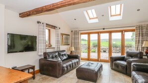 A spacious living room with a brown leather sofa, a wooden table, and a television mounted on the wall. There are two windows with curtains, a large sliding glass door leading to a garden, and a wooden ceiling beam. - Rose Hips Barn (Chorley)