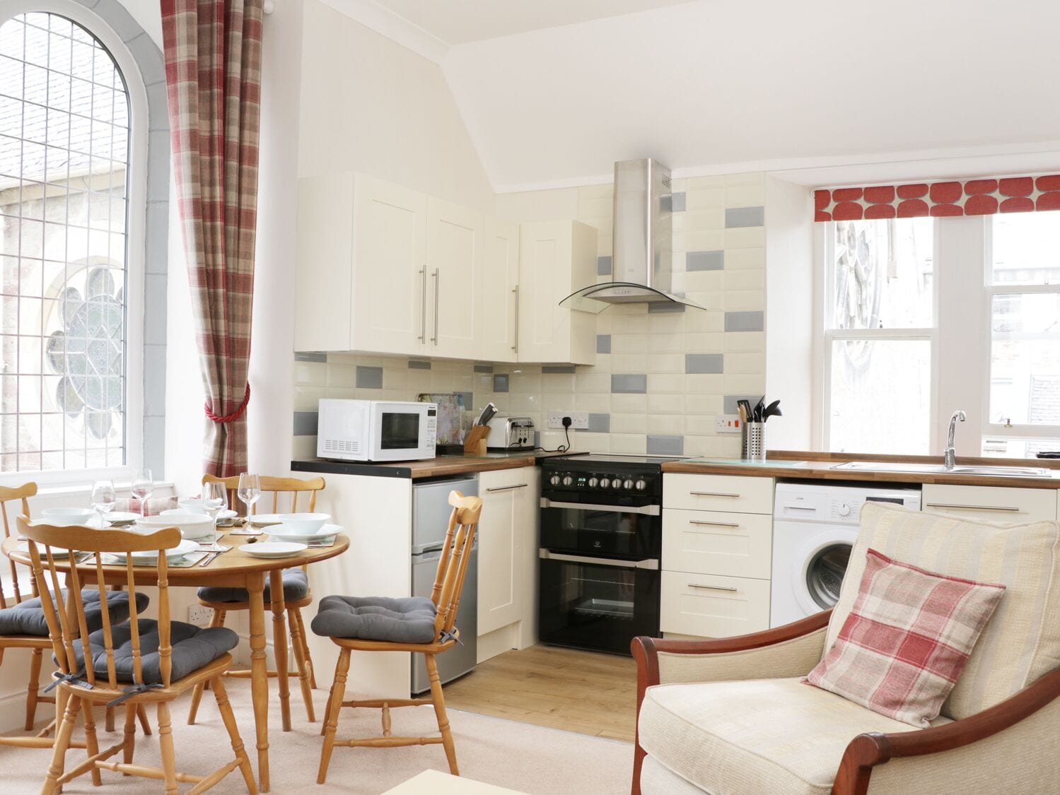 A modern kitchen with white cabinets, a microwave, a stove, and a sink. There is a dining table with chairs and a sofa with a plaid pillow.