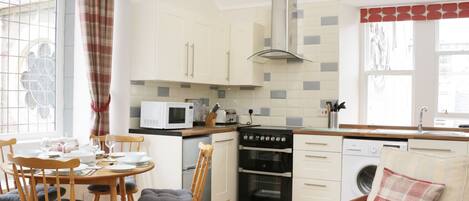 A modern kitchen with white cabinets, a microwave, a stove, and a sink. There is a dining table with chairs and a sofa with a plaid pillow.