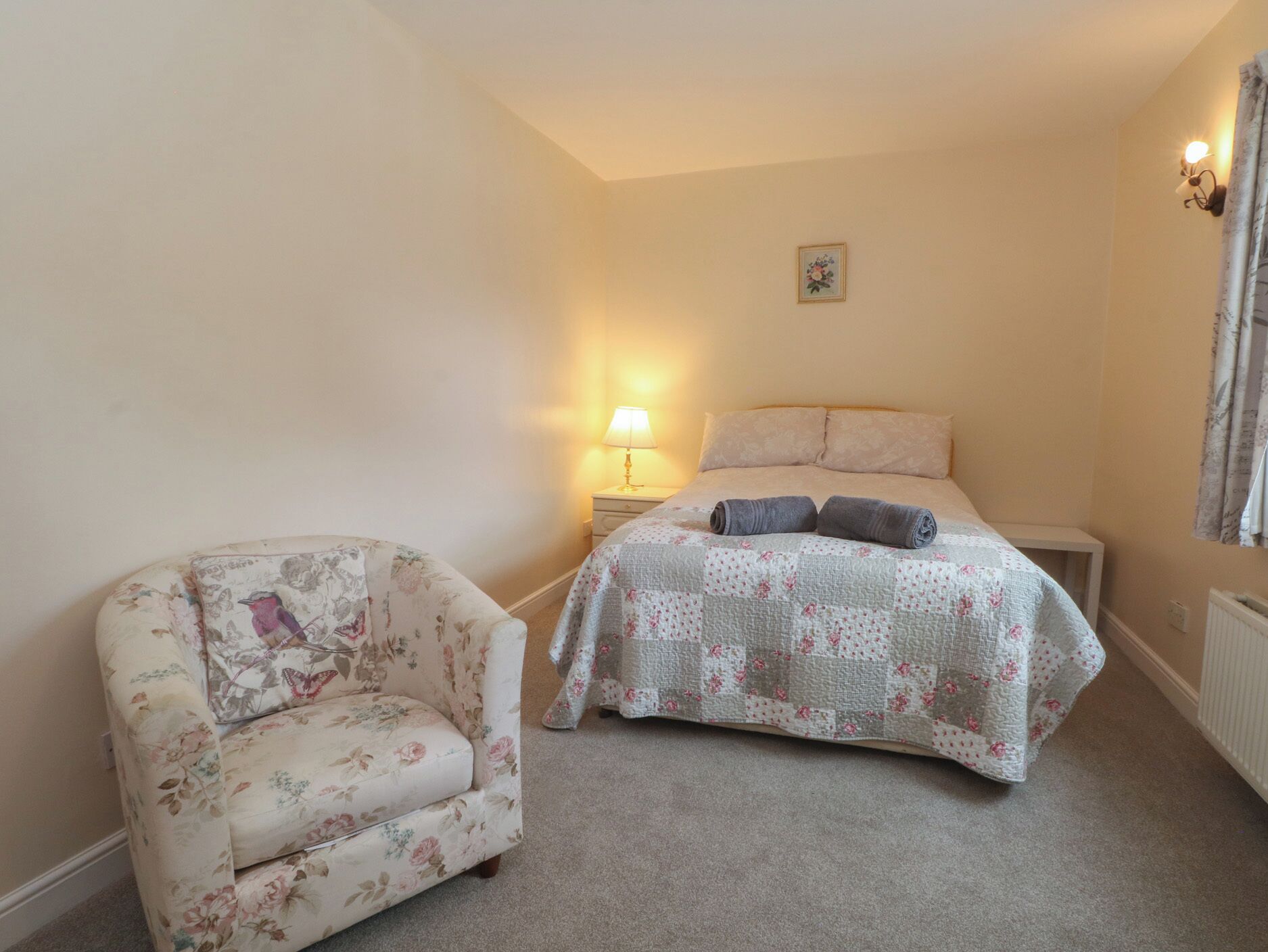 A bedroom with a bed, a nightstand, a lamp, and a floral armchair.