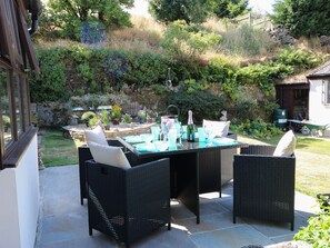 A patio with a glass-top table and wicker chairs, surrounded by a garden with various plants and a stone wall. - West End House (Bradford)