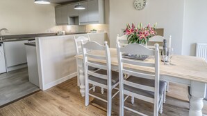 A modern kitchen with white cabinetry, a dining area with white chairs, a wooden table, and a clock on the wall. - Y Cilgwyn (Blaenau Ffestiniog)