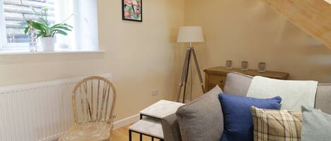 A cozy living room with a wooden chair, a sofa with cushions, a floor lamp, and a window with a plant.