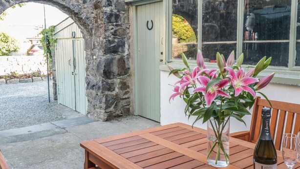 A wooden table with a bottle of wine and glasses, a vase of pink lilies, and a stone archway leading to a door. - Y Cilgwyn (Blaenau Ffestiniog)