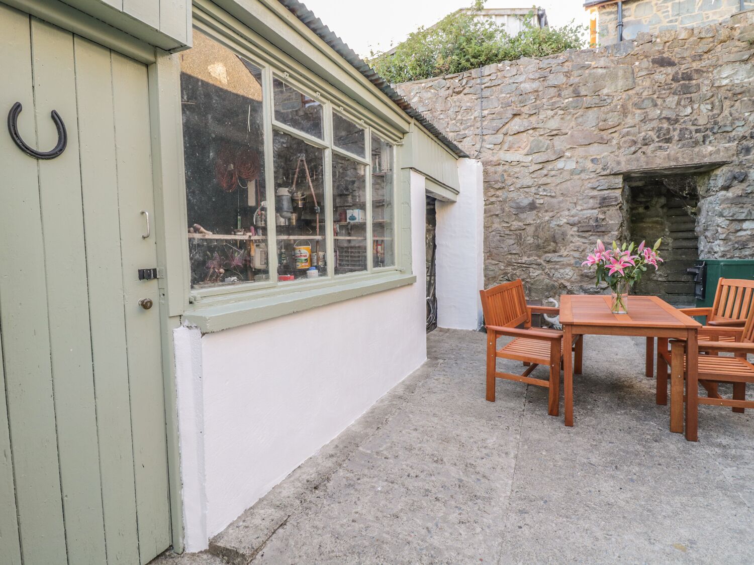 A stone wall adjacent to a building with a window displaying various items. There is a table with chairs and a vase of flowers.