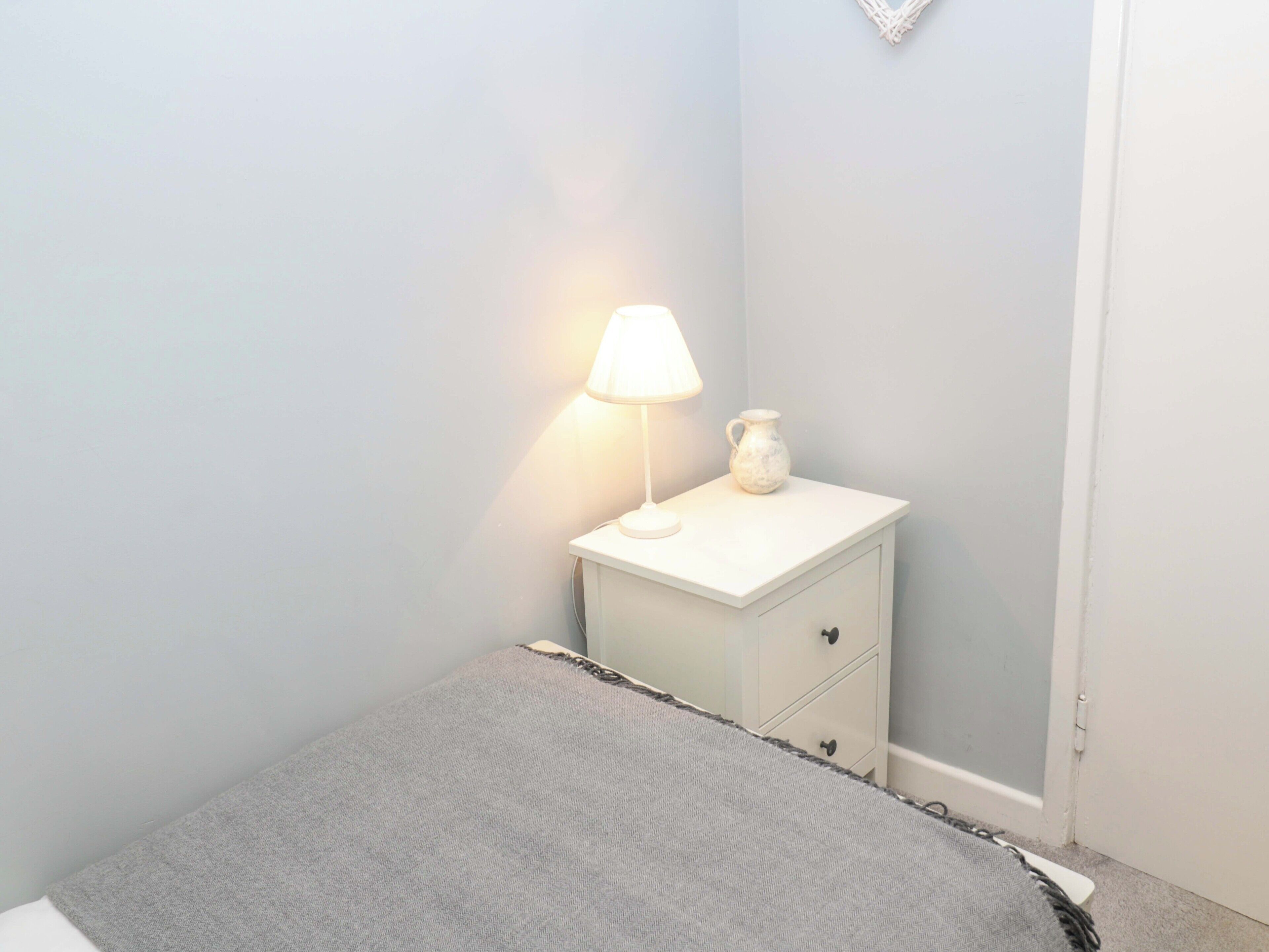 A bedroom with a bed, a nightstand with a lamp and a vase, and a door.