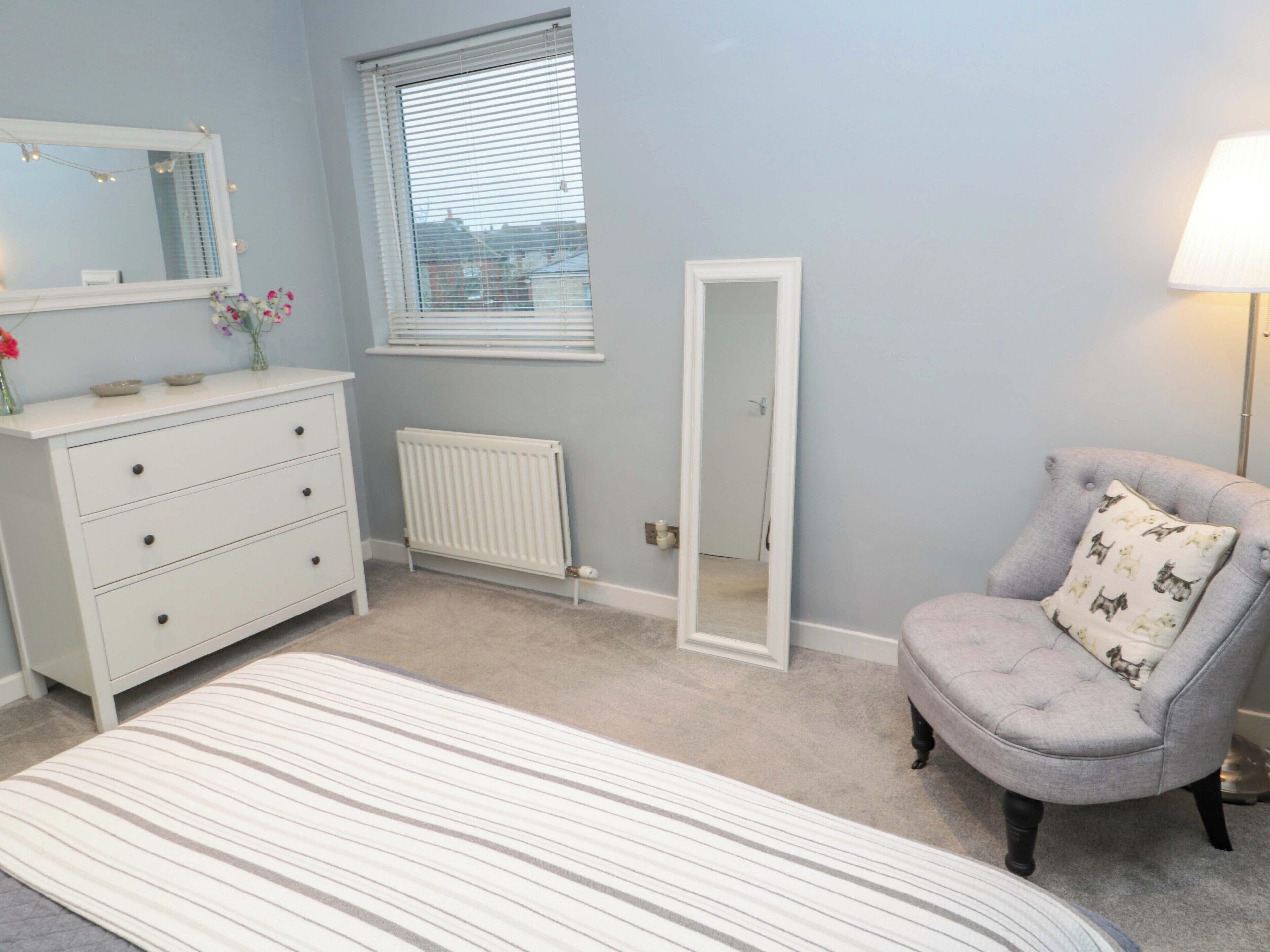 Ein Schlafzimmer mit Bett, Schminktisch, Stuhl, Heizkörper, Fenster mit Jalousien und Spiegel.
