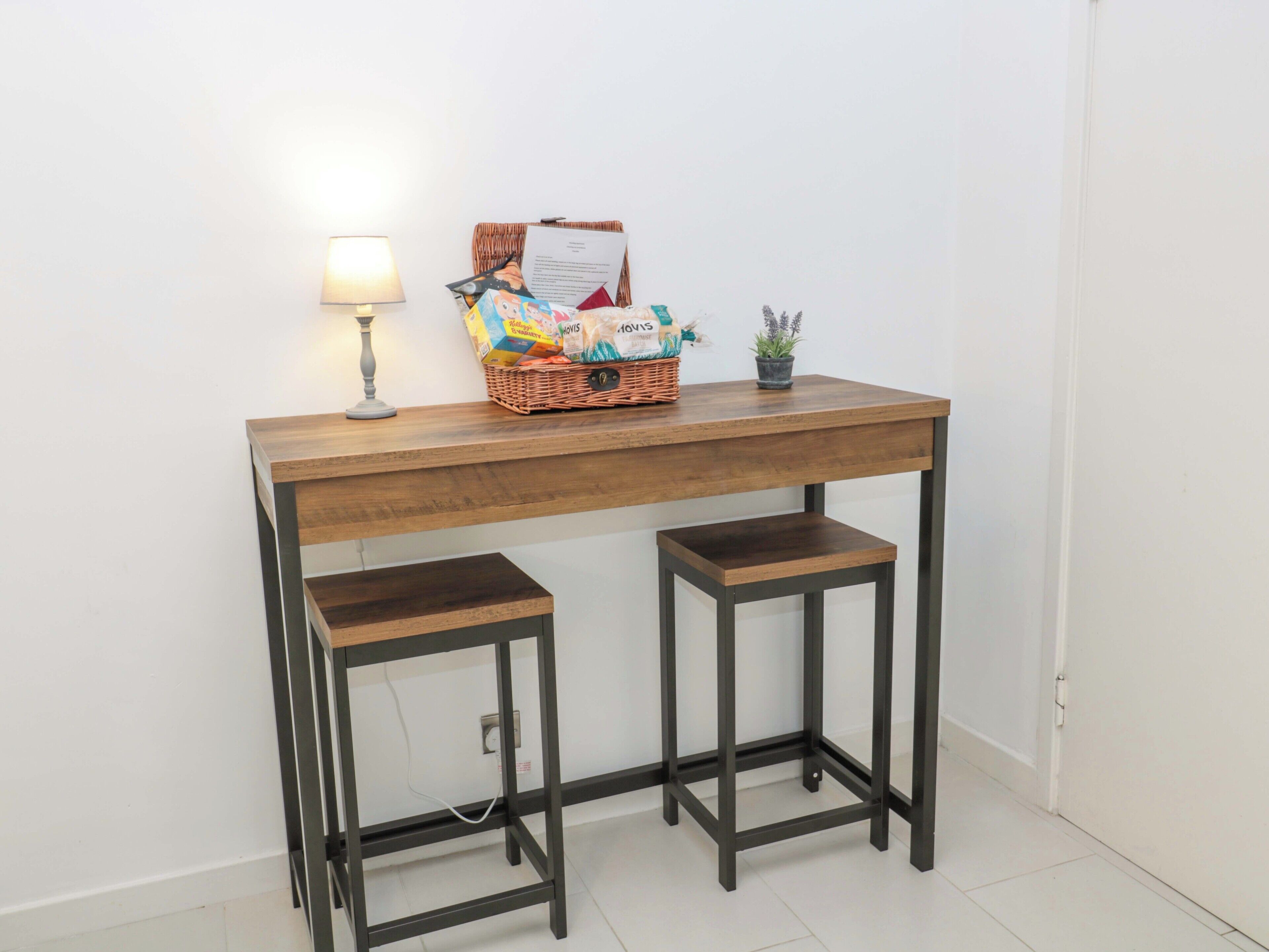 A wooden desk with two attached stools, a lamp, a basket with items, and a small plant.