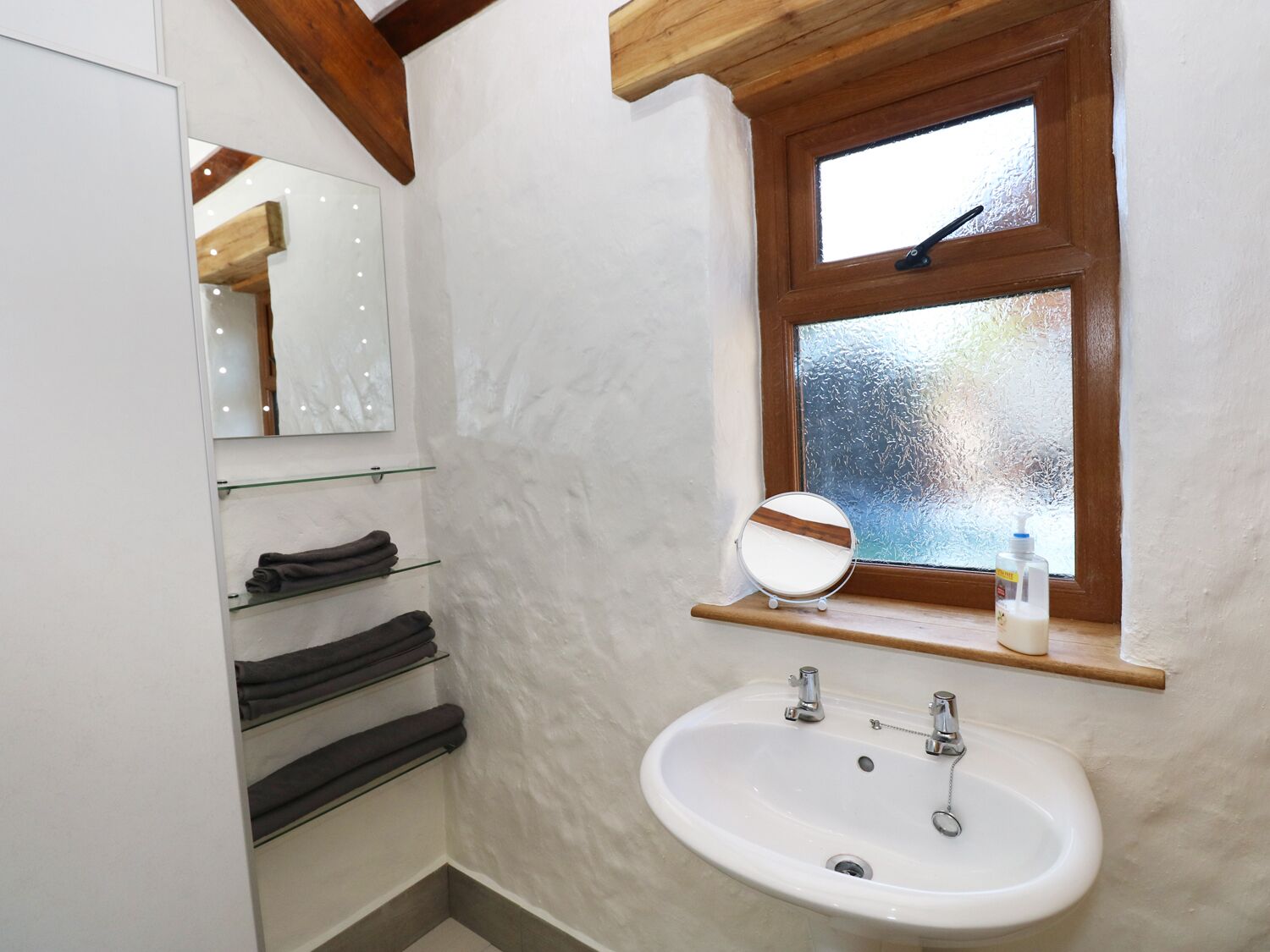 A bathroom with a white sink, a mirror, a window with frosted glass, and a shelf with towels.